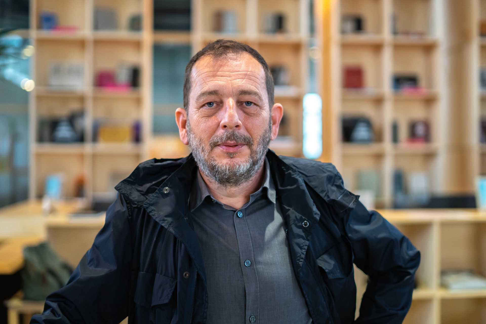 Jeton Jagxhiu is standing in a room in the Autostrada Hangar in front of a shelve with many items in it. Jeton smiles into the camera and wears a black jacket.