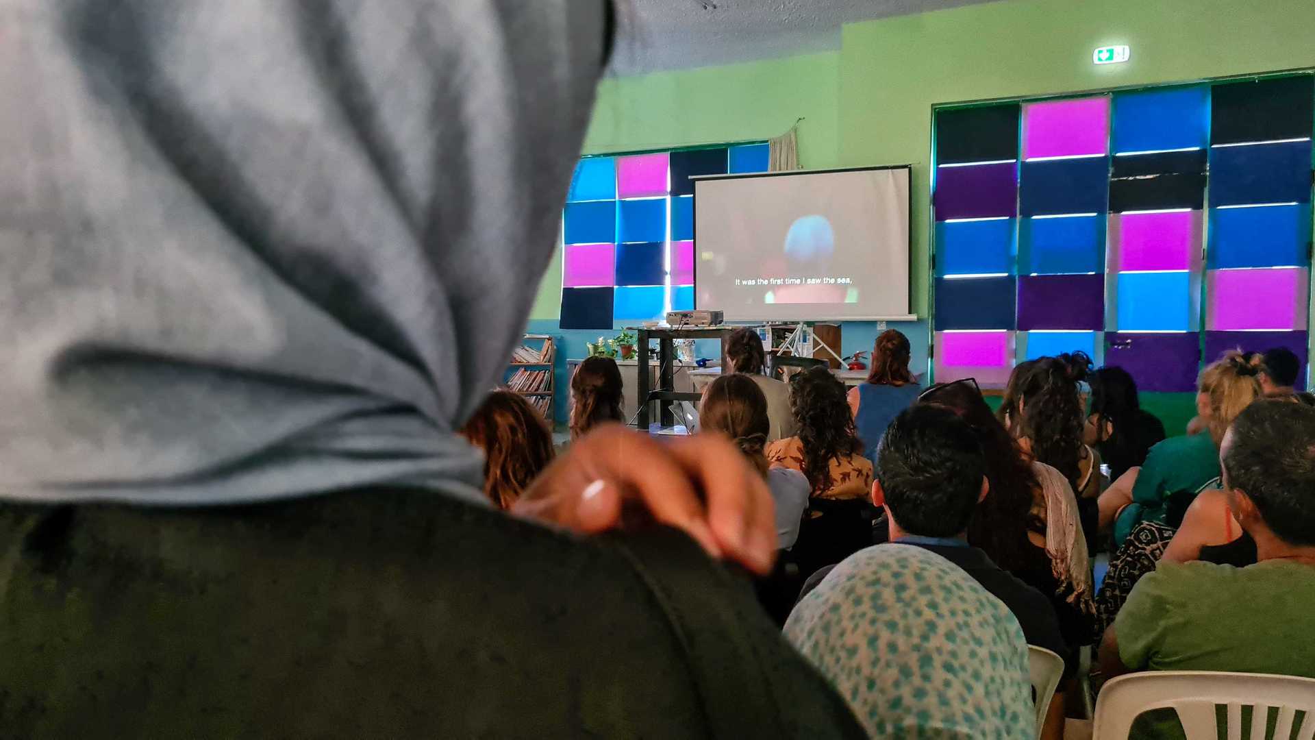 A very crowded room. People are looking to a big screen, where a movie is shown.