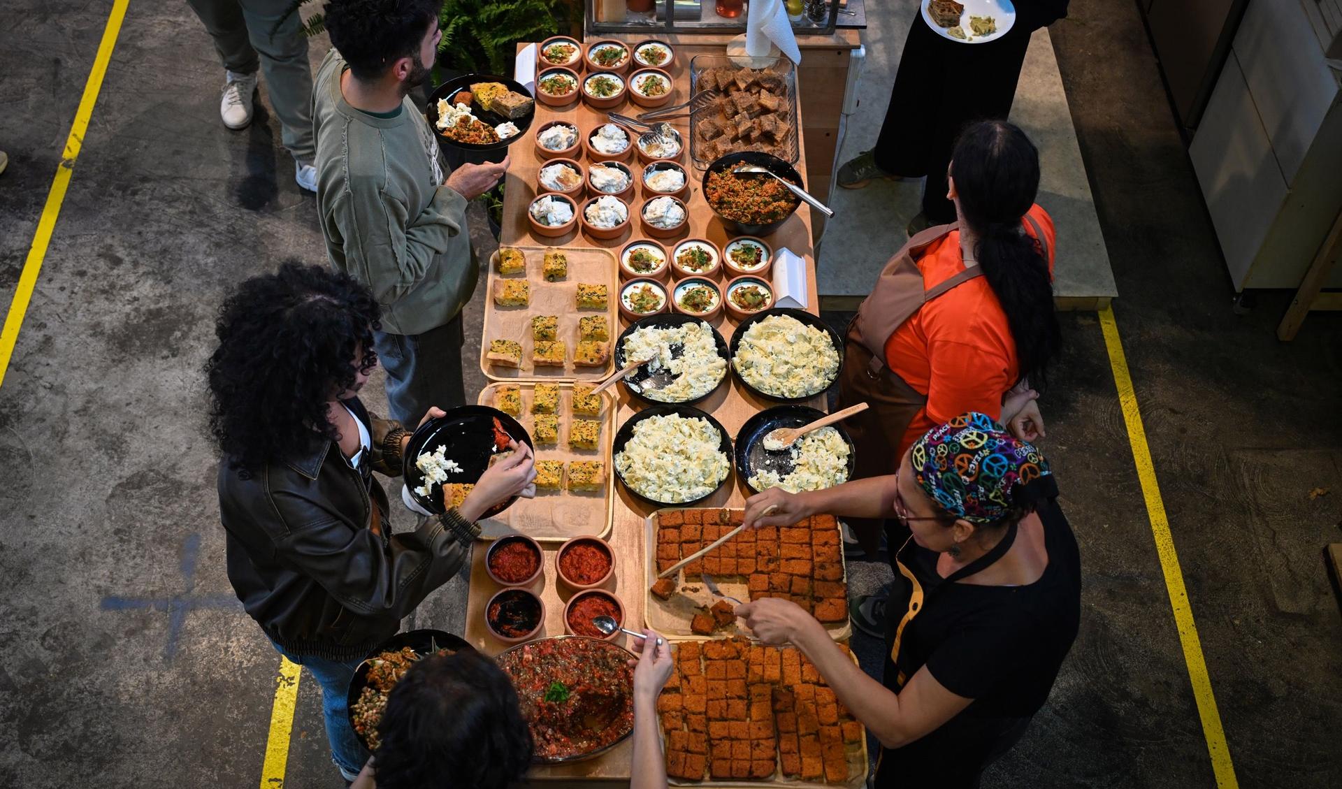 Many people are standing at a table. Some are getting food, others are serving food.