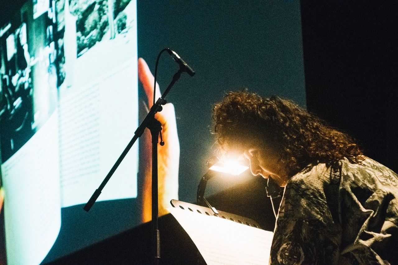 A person is sitting on the right and is playing the clarinette and reading her notes. A small light is shining. In the background a video projection is running. It is a concert.