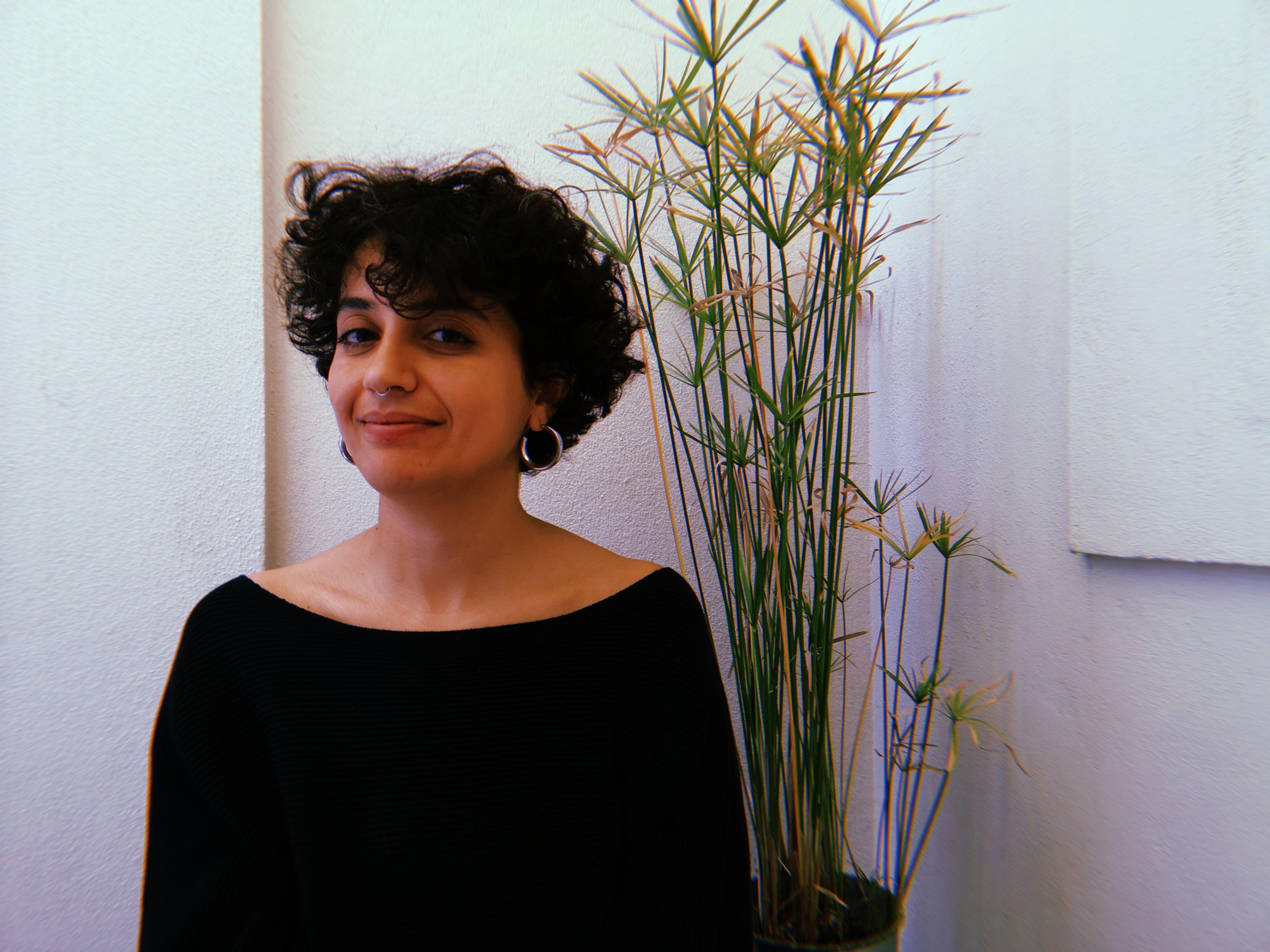 Aylin Cankaya is standing in white room with a plant in the background. Aylin is smiling into the camera, wearing earrings. Aylin has short curly hair.