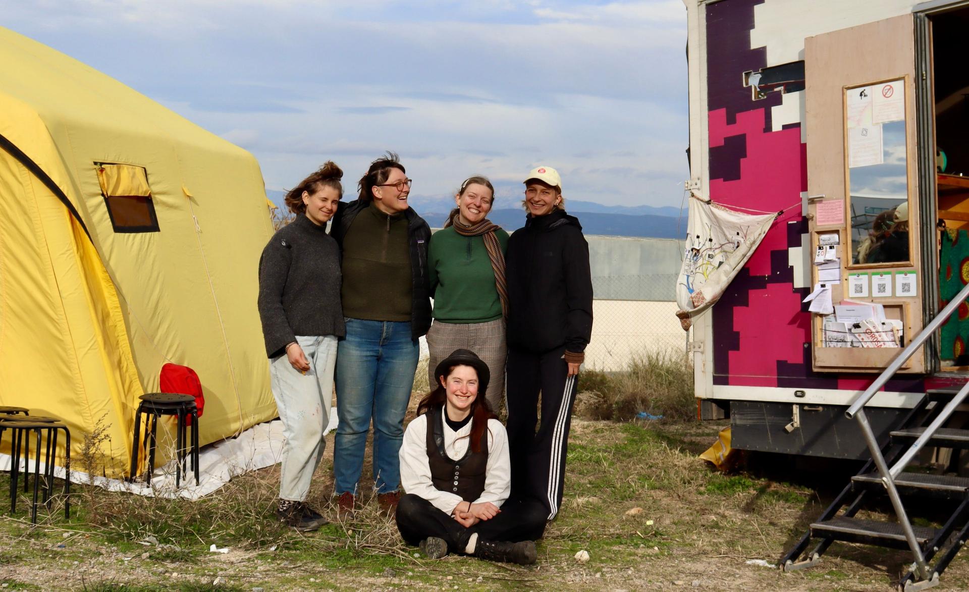 Five person are standing between a tent and a truck next to a refugee camp