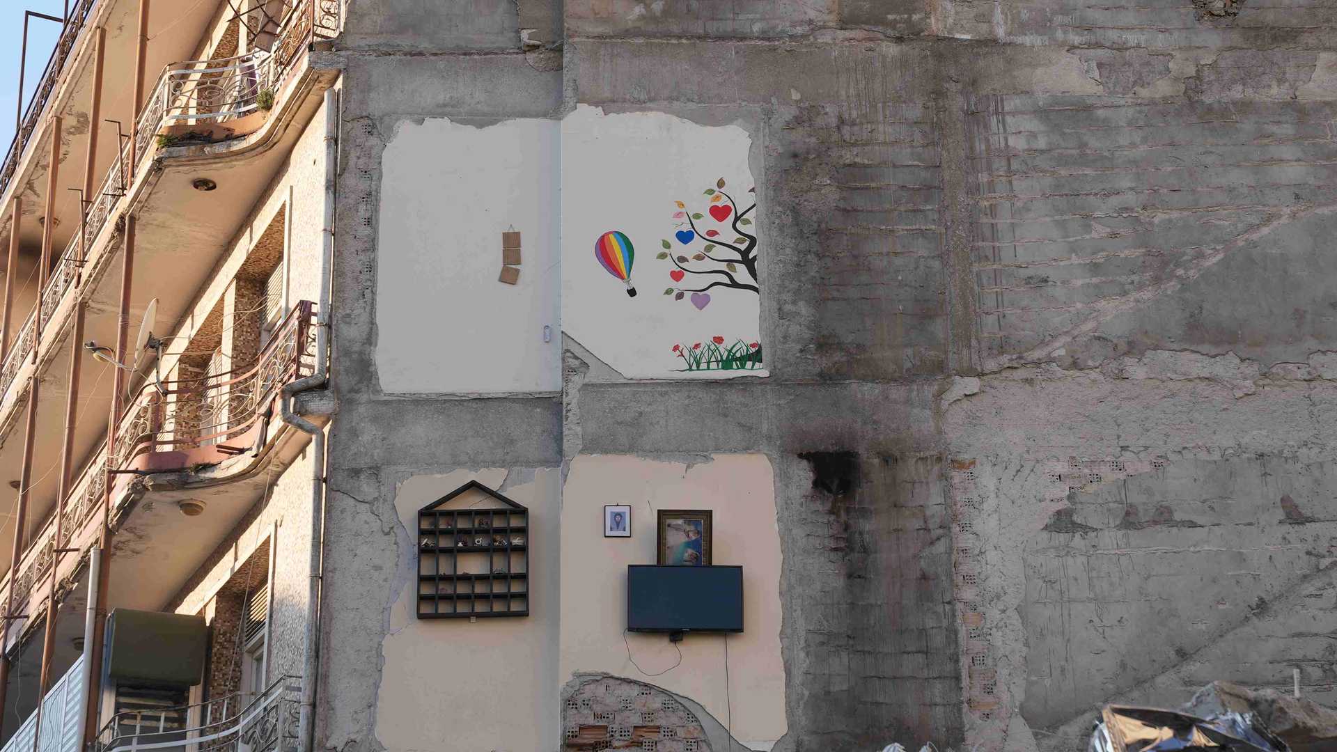 A wall of a demolished house. There are leftovers of the walls of the rooms, one where still a TV-screen is attached to the wall, another one with a shelf.