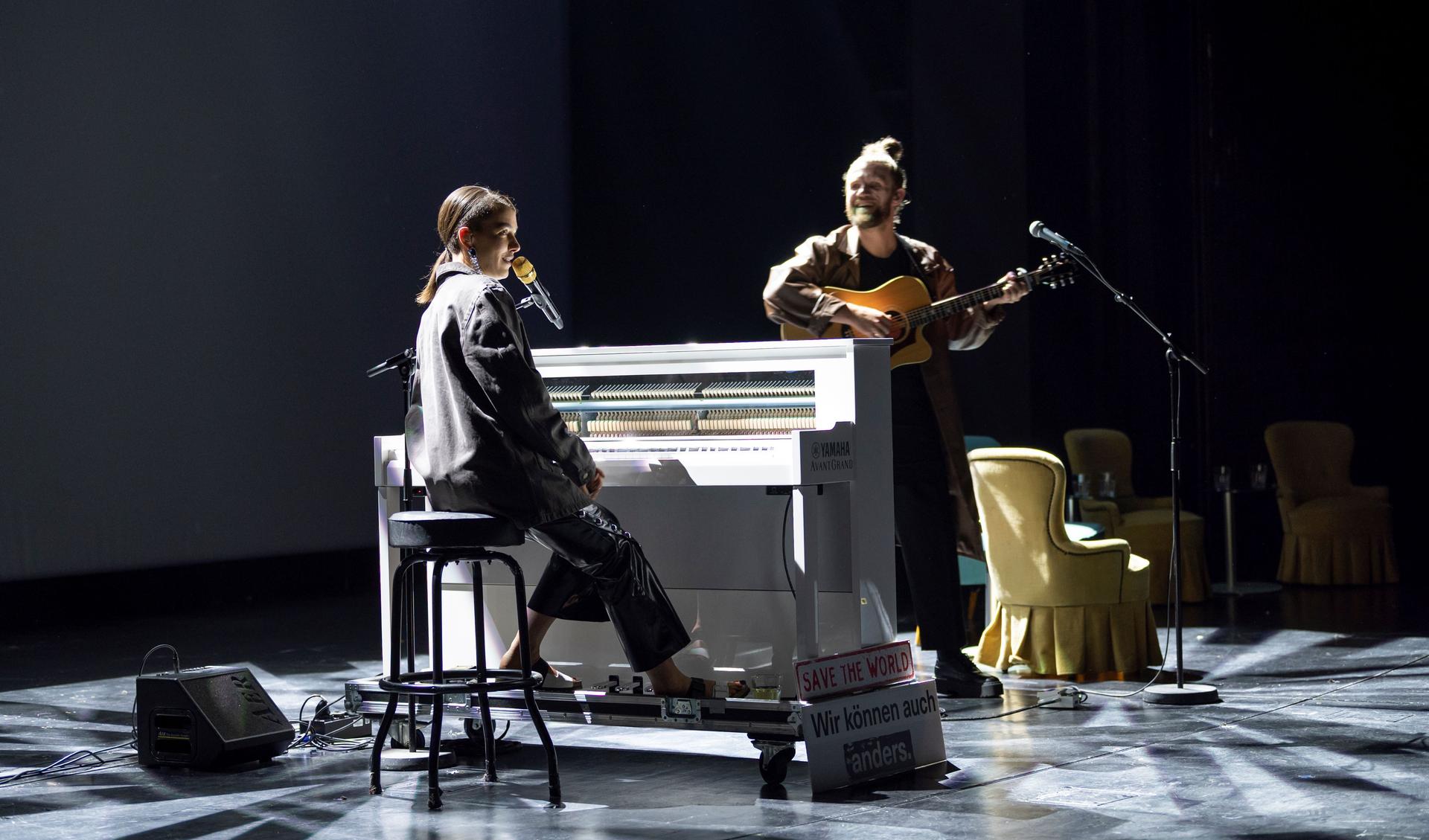 A women is sitting at a piano and is playing it. Next to her is a guy playing a guitar and smiling at her. The two are on a stage.