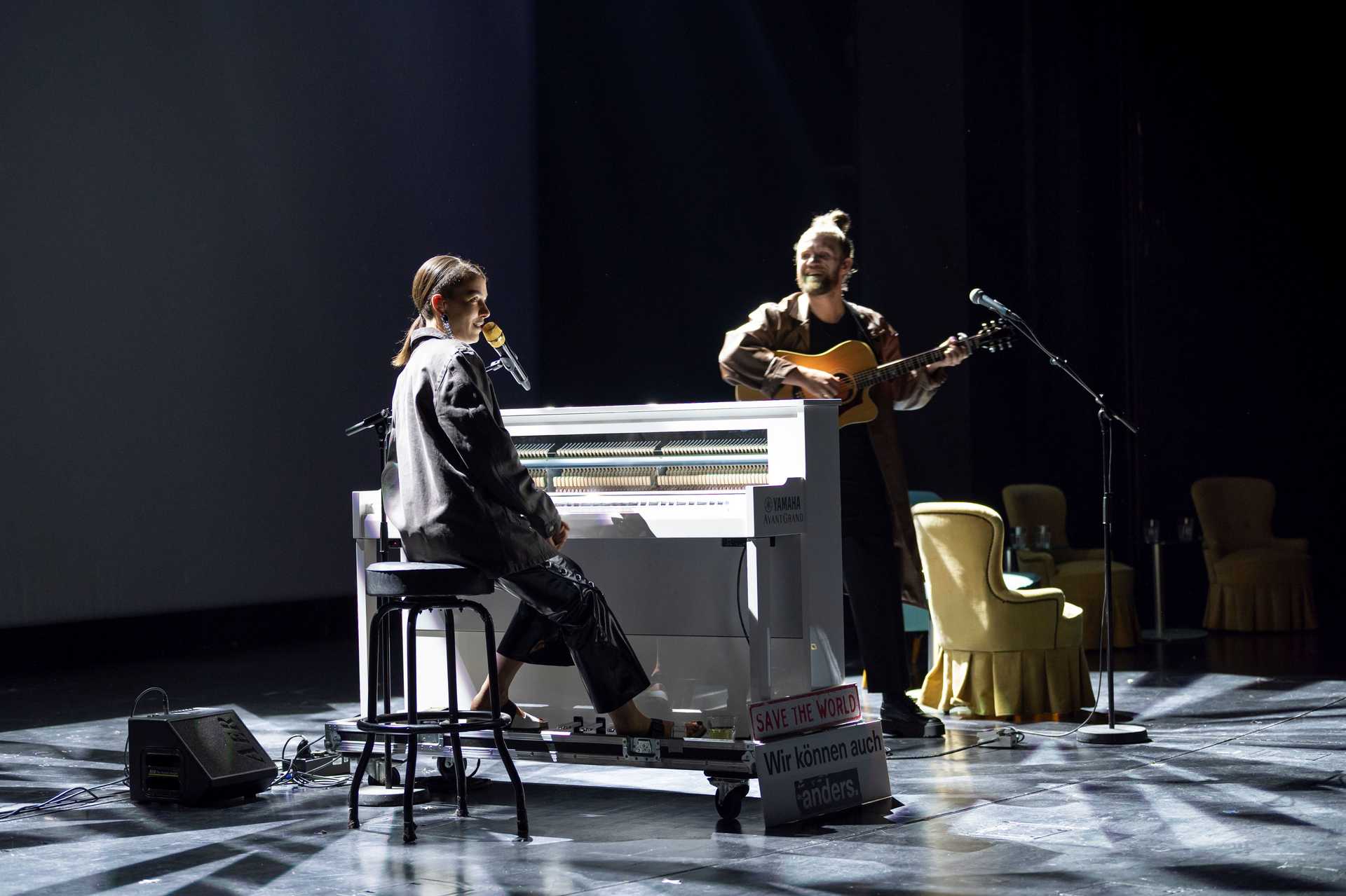 A women is sitting at a piano and is playing it. Next to her is a guy playing a guitar and smiling at her. The two are on a stage.