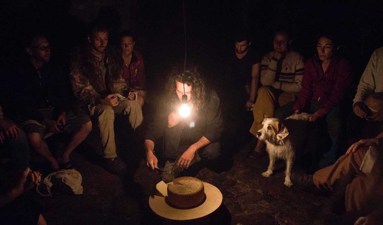 People are sitting around a cheese. One person sits in the middle and cuts the cheese. There is one dimmed light shining.