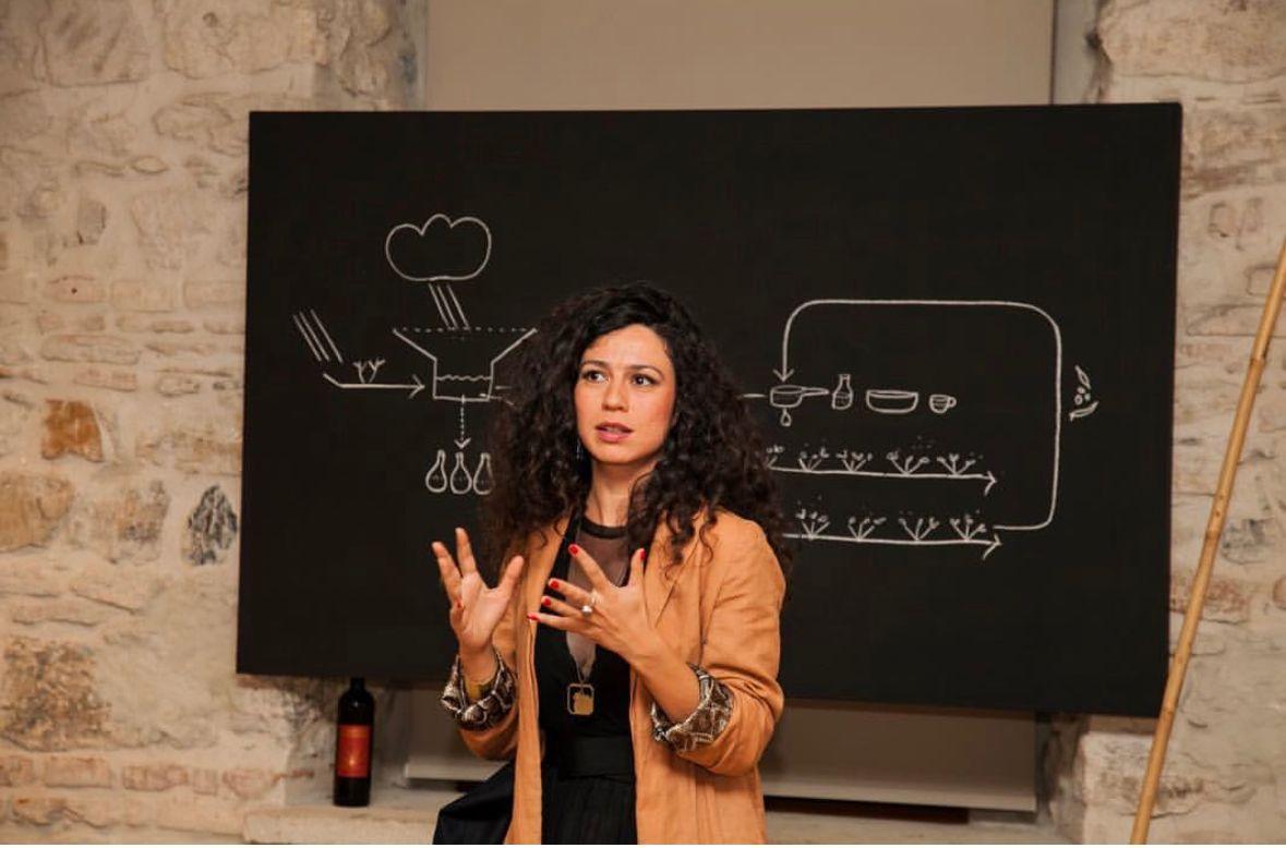 Sinem Dişli stands in a room in front of a blackboard with notes on it. They seem to sketch a weather phenomena. Sinem is gesturing in front of the blackboard, wearing a light jacket and havin long curled hair.