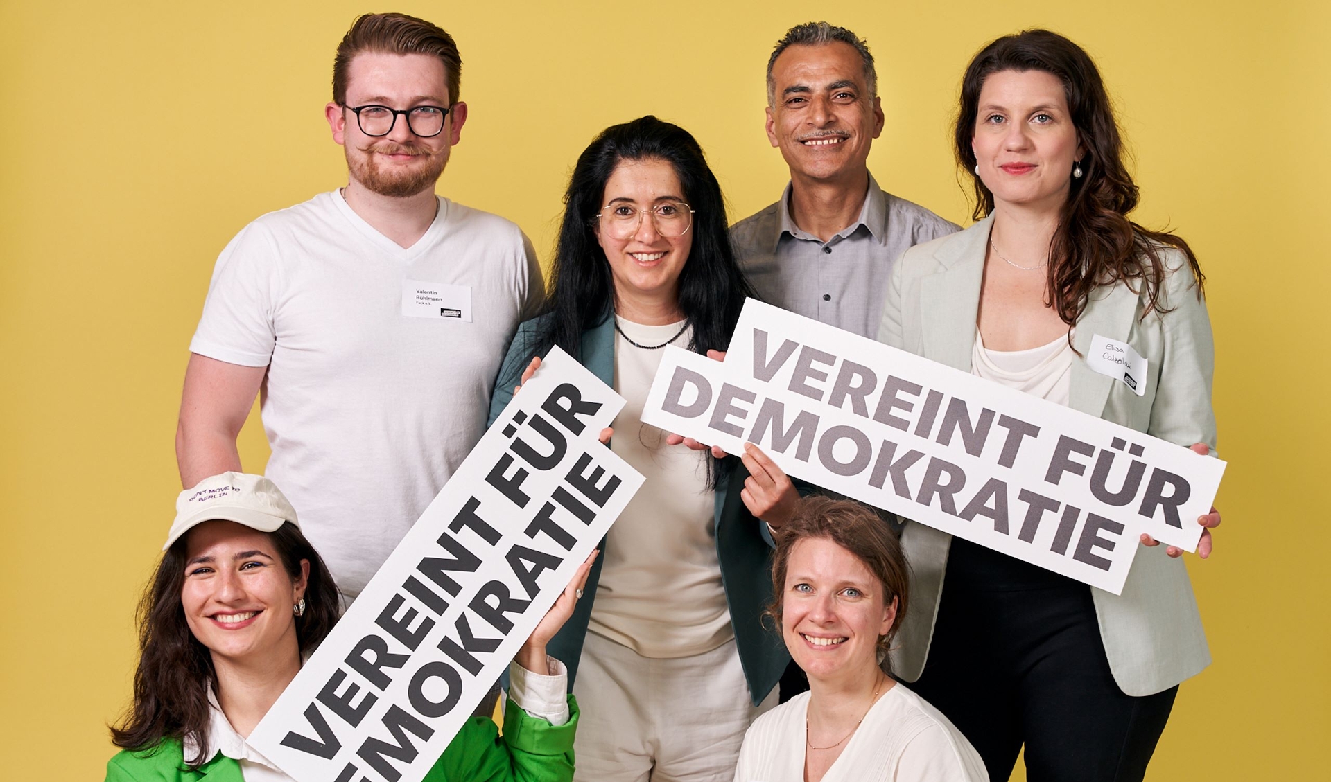six people in front of a yellow background are holding signs saying "United for Democray"