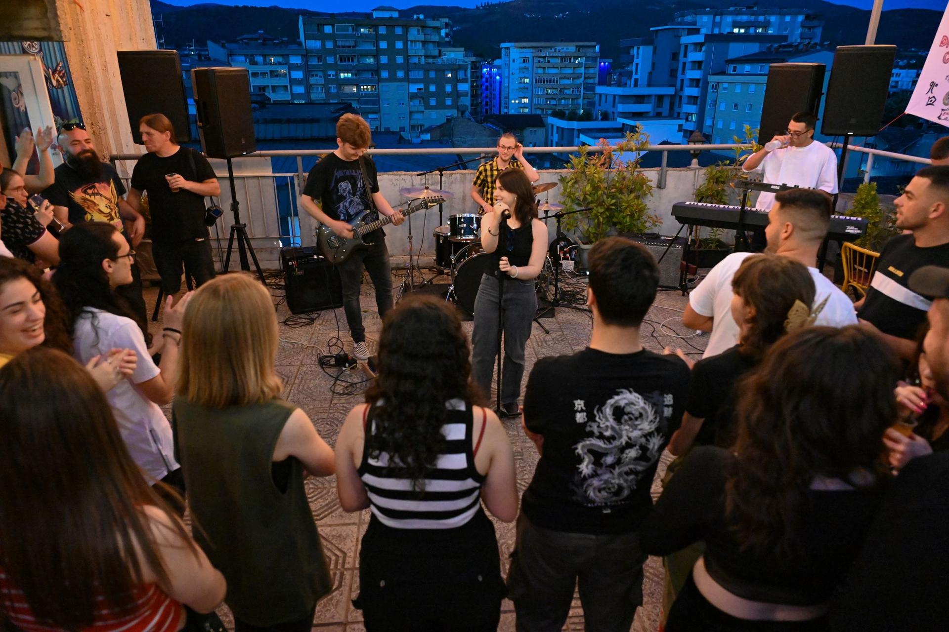 A crowd is standing on a terrace and is listening to a band of three people performing music