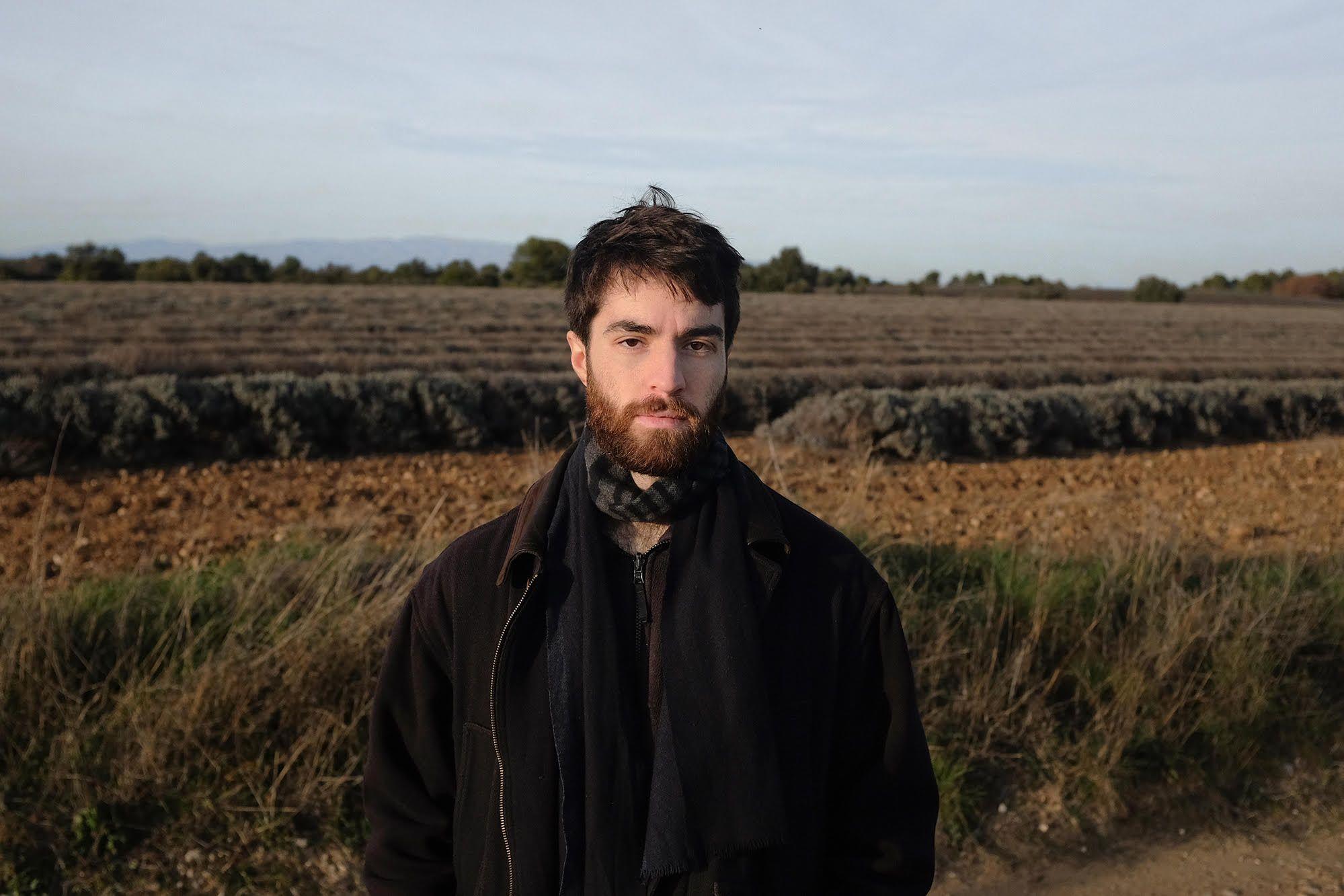 A picture of a man standing in a field. He has dark hair and a beard, he wears a scarve and a coat. The sky is cloudy.