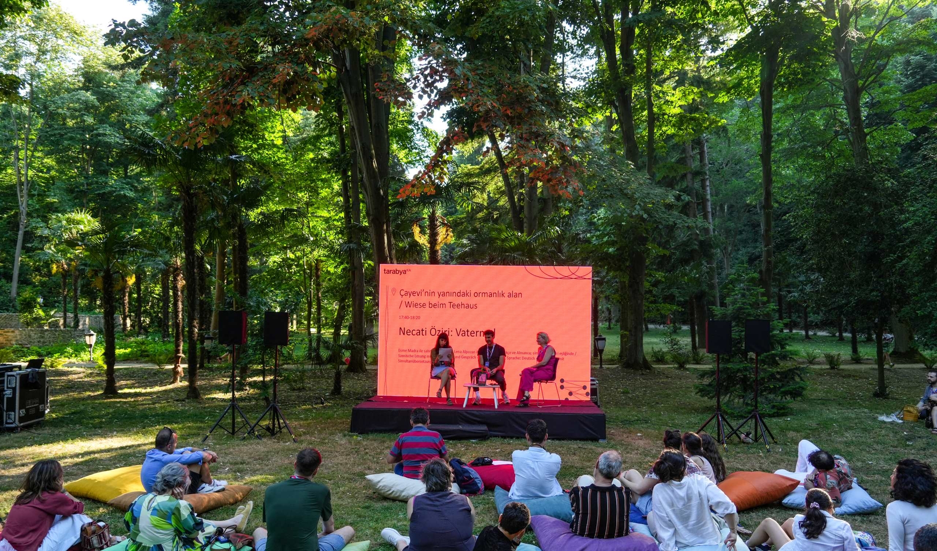 Three people are sitting on a stage in a huge garden full of trees. Many people are hanging out in front of the stage and are listening to the talk.