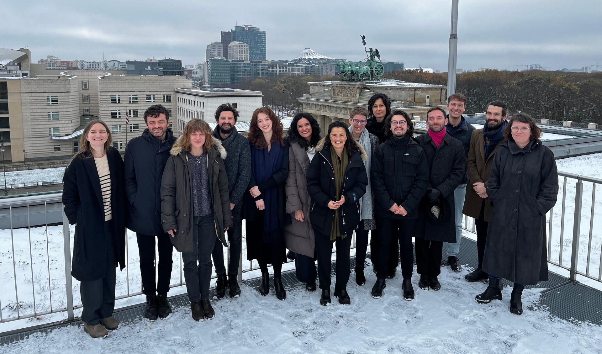 The team of the Hubs of the Allianz Foundation are standing on the roof top terrace of a building. It is winter, it is grey and in the background you see the skyline of Berlin