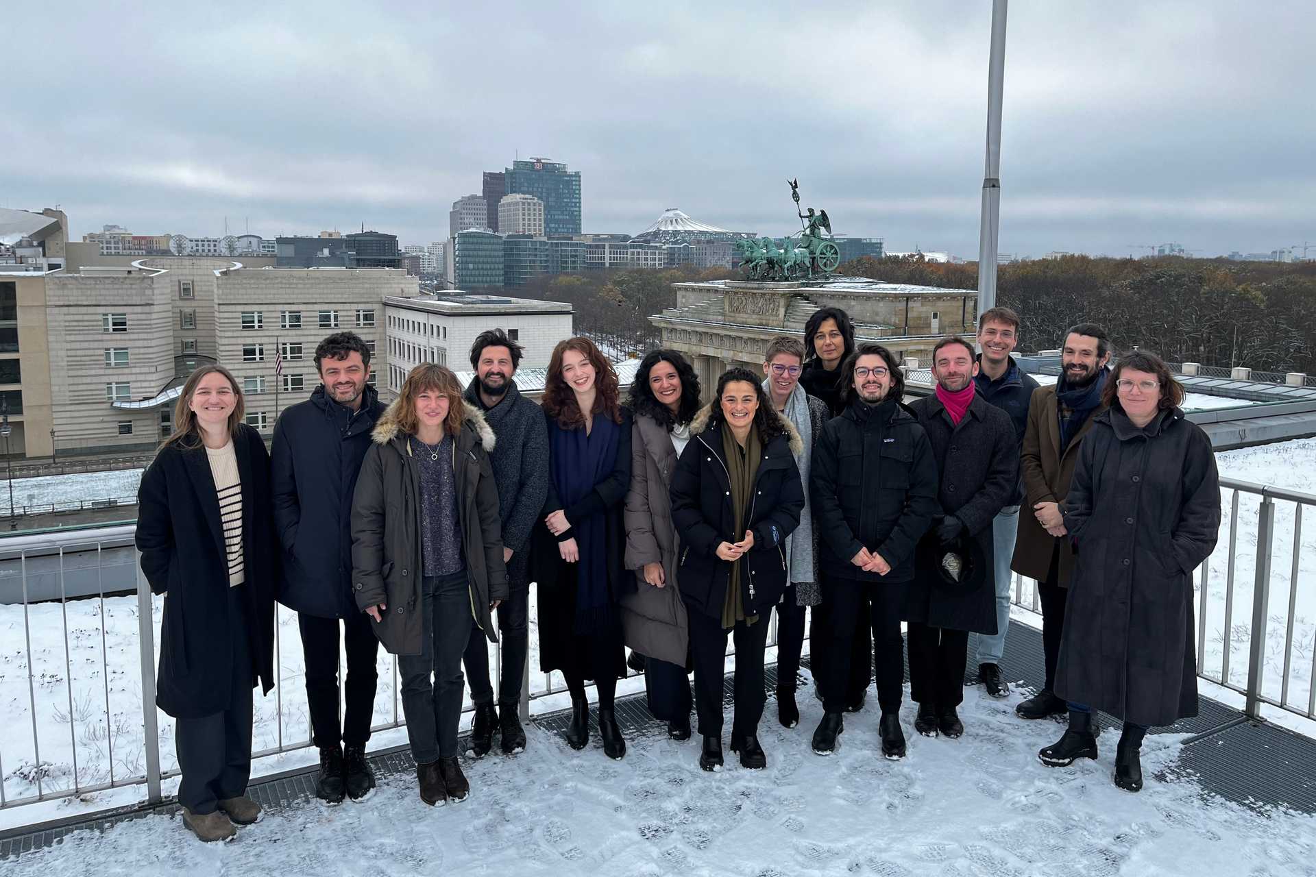 The team of the Hubs of the Allianz Foundation are standing on the roof top terrace of a building. It is winter, it is grey and in the background you see the skyline of Berlin