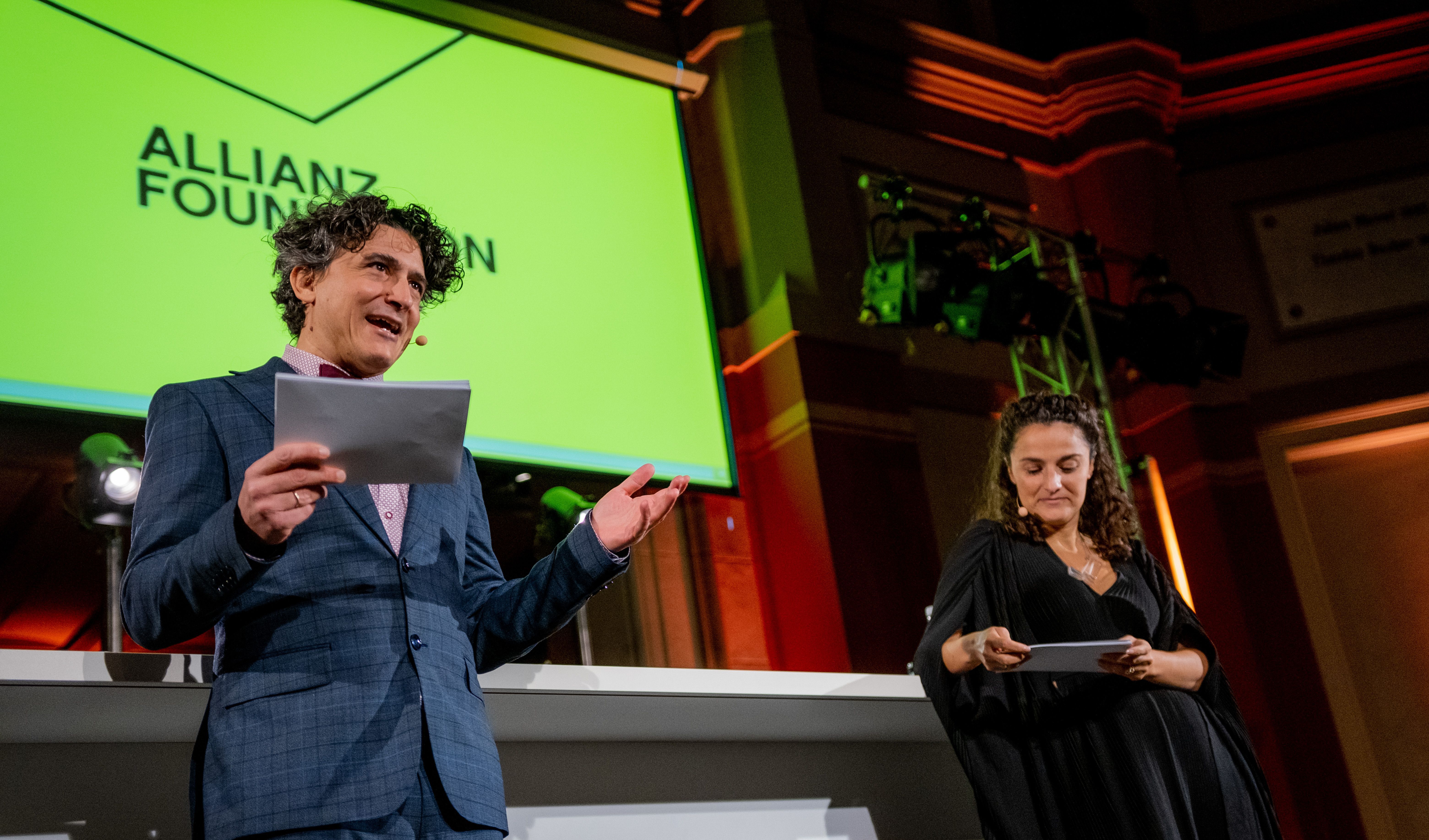 Conductor and Allianz Foundation Partner Lefteris Veniadis stands next to Esra Kücük, Allianz Foundation CEO on a stage. They both wear very elegant clothes and are hosting the evening