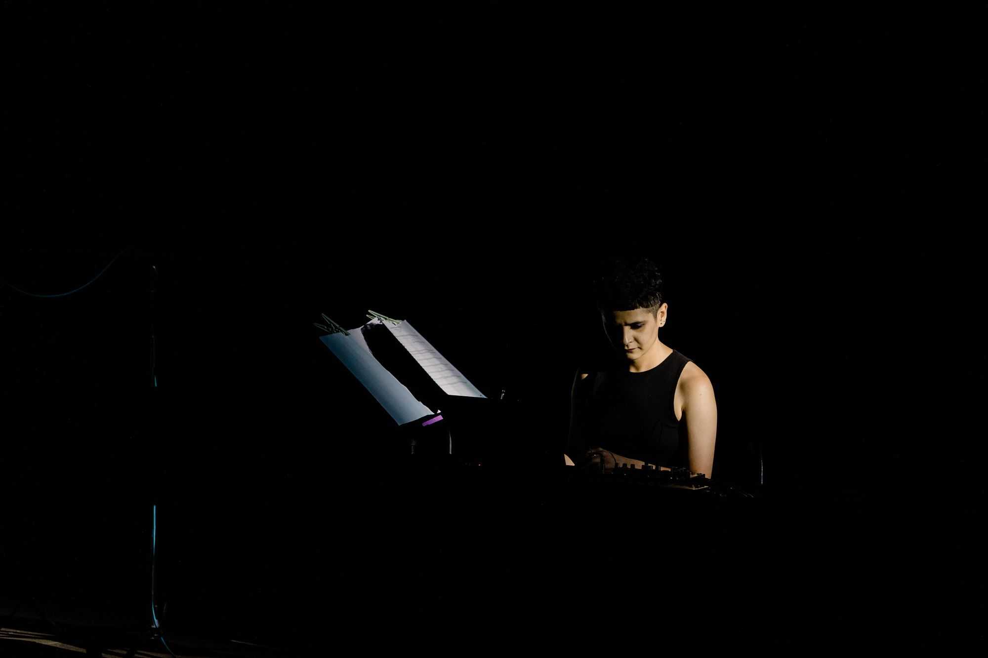 A women is sitting at a piano and is playing it. Around her it's pitch black.