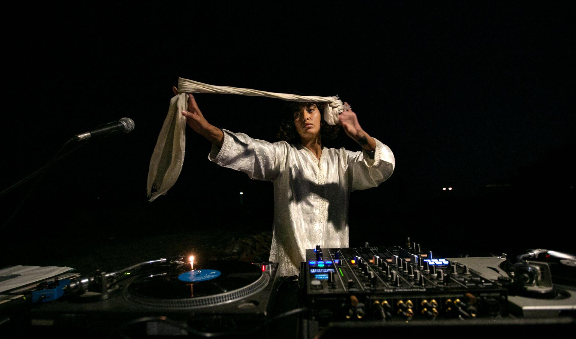 The artist Leila Bencharnia is playing at DJdesks in the nighttime and holds a scarve in her hands