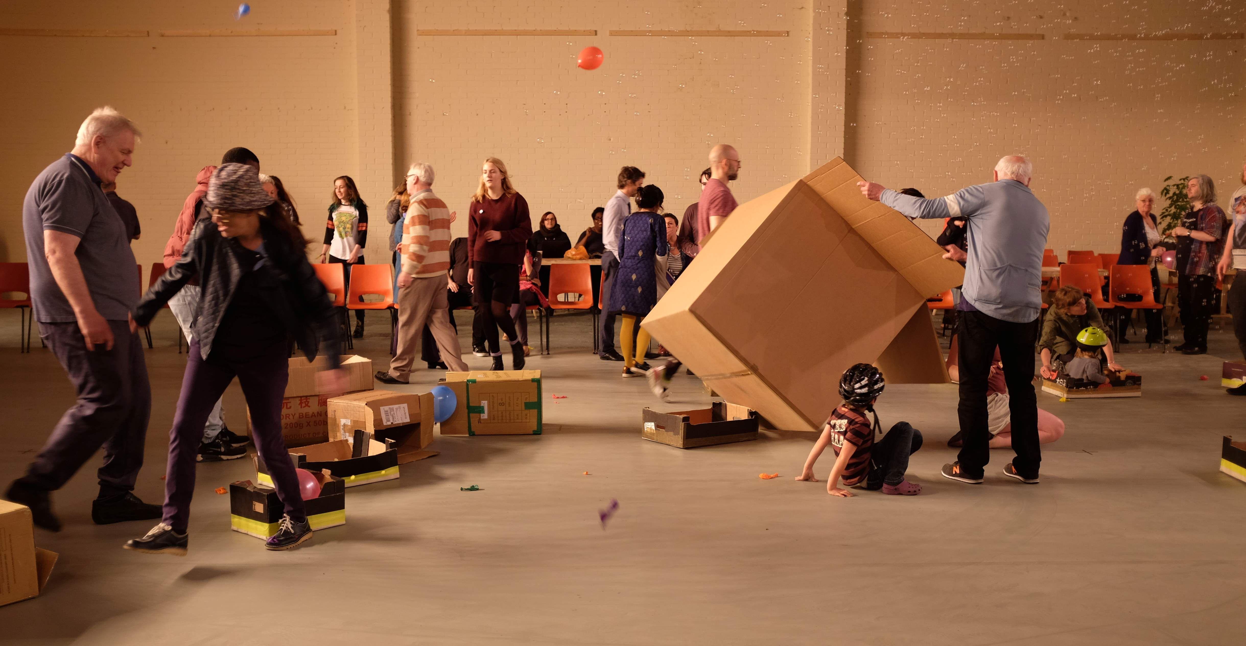 Many people are assembled in a hall. They are interacting and handling cardboard boxes