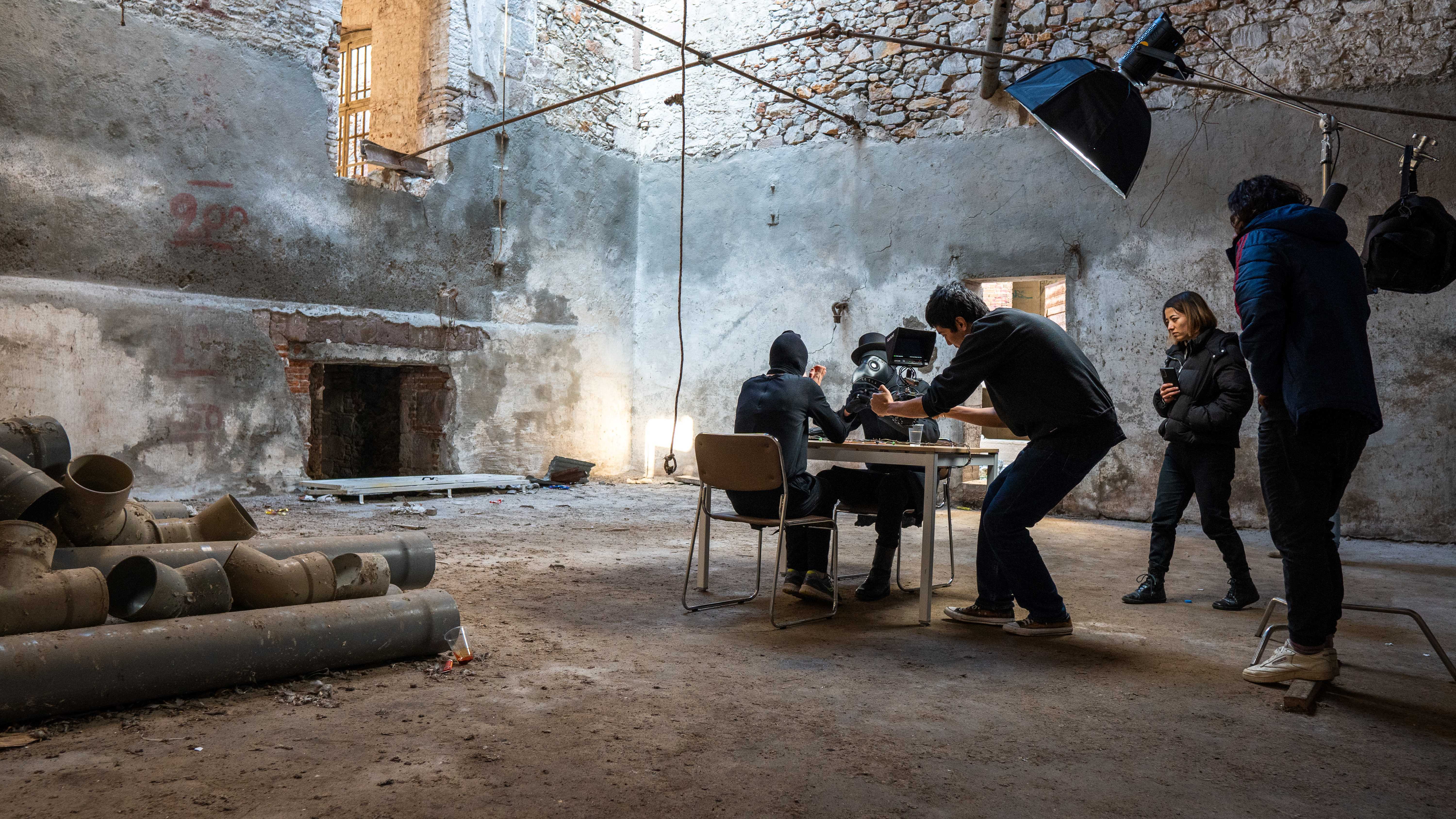 Five persons in a room are making a movie. Two are sitting at a table in a shabby and cold room and the others are filming them.