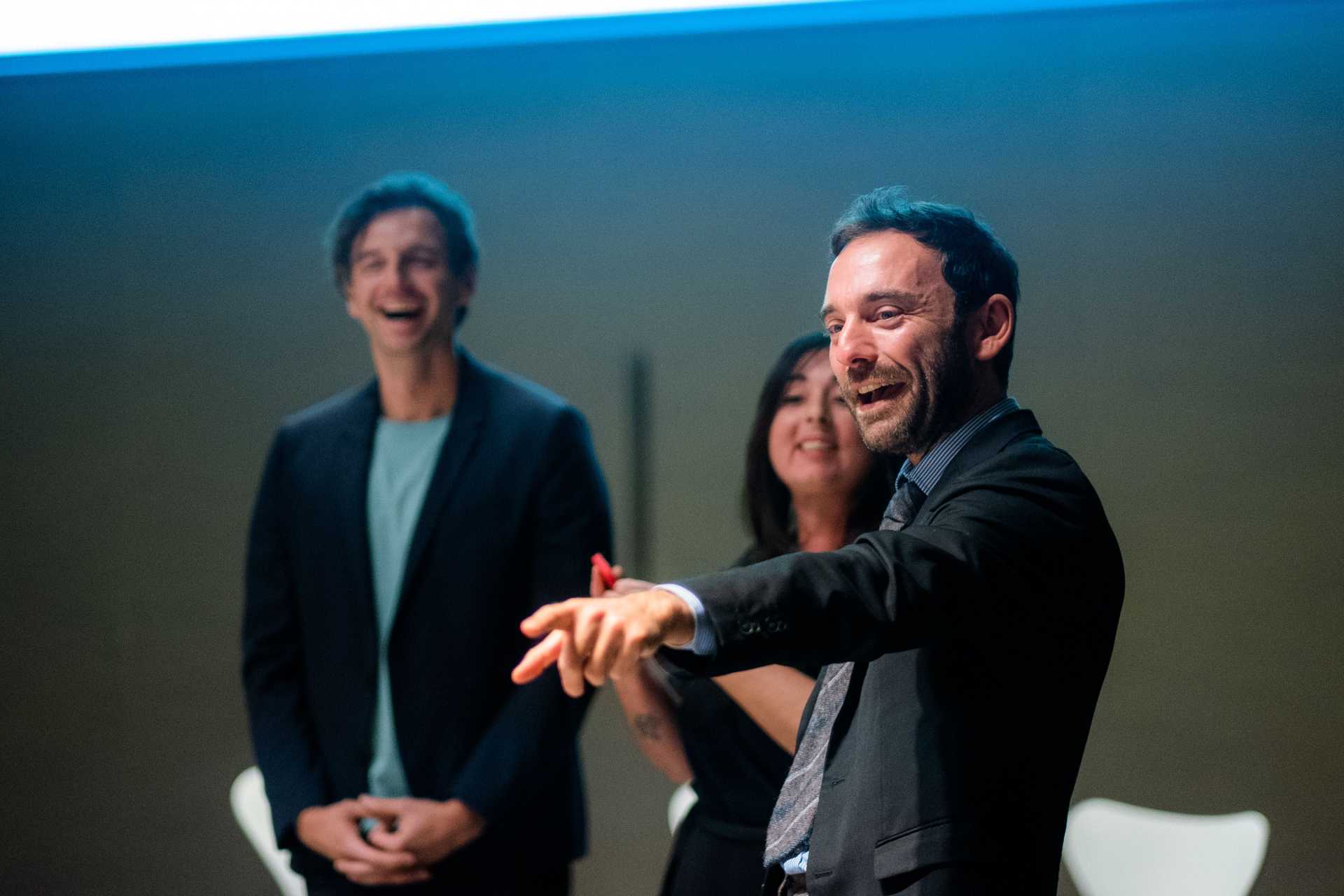 Three persons are standing in a room, two are laughing, while a tghird person is adressing the guests with something that makes them laugh.