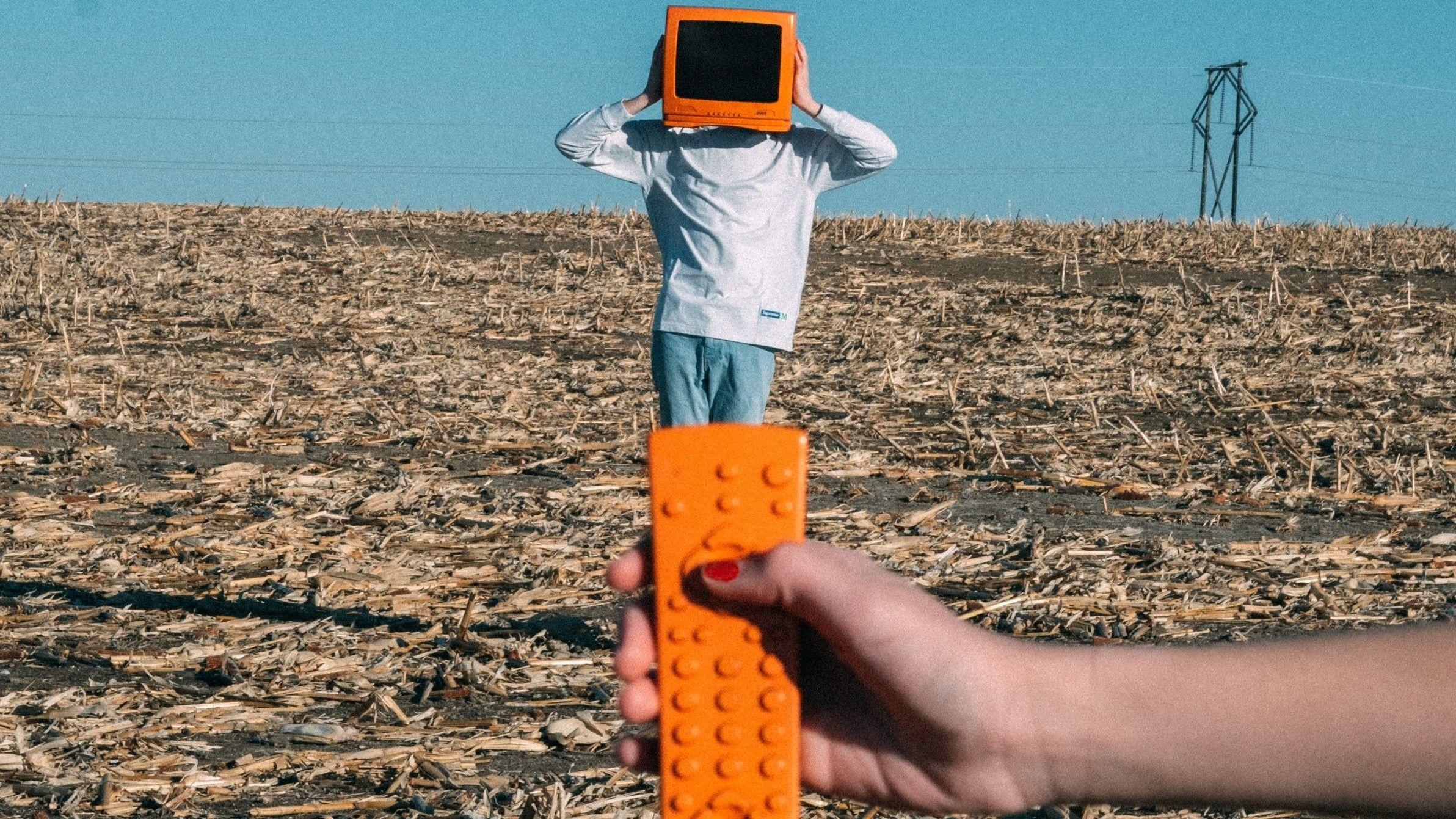 A person is standing on a harvested crops field and wears a TV on his head. A hand holds an orange remote control