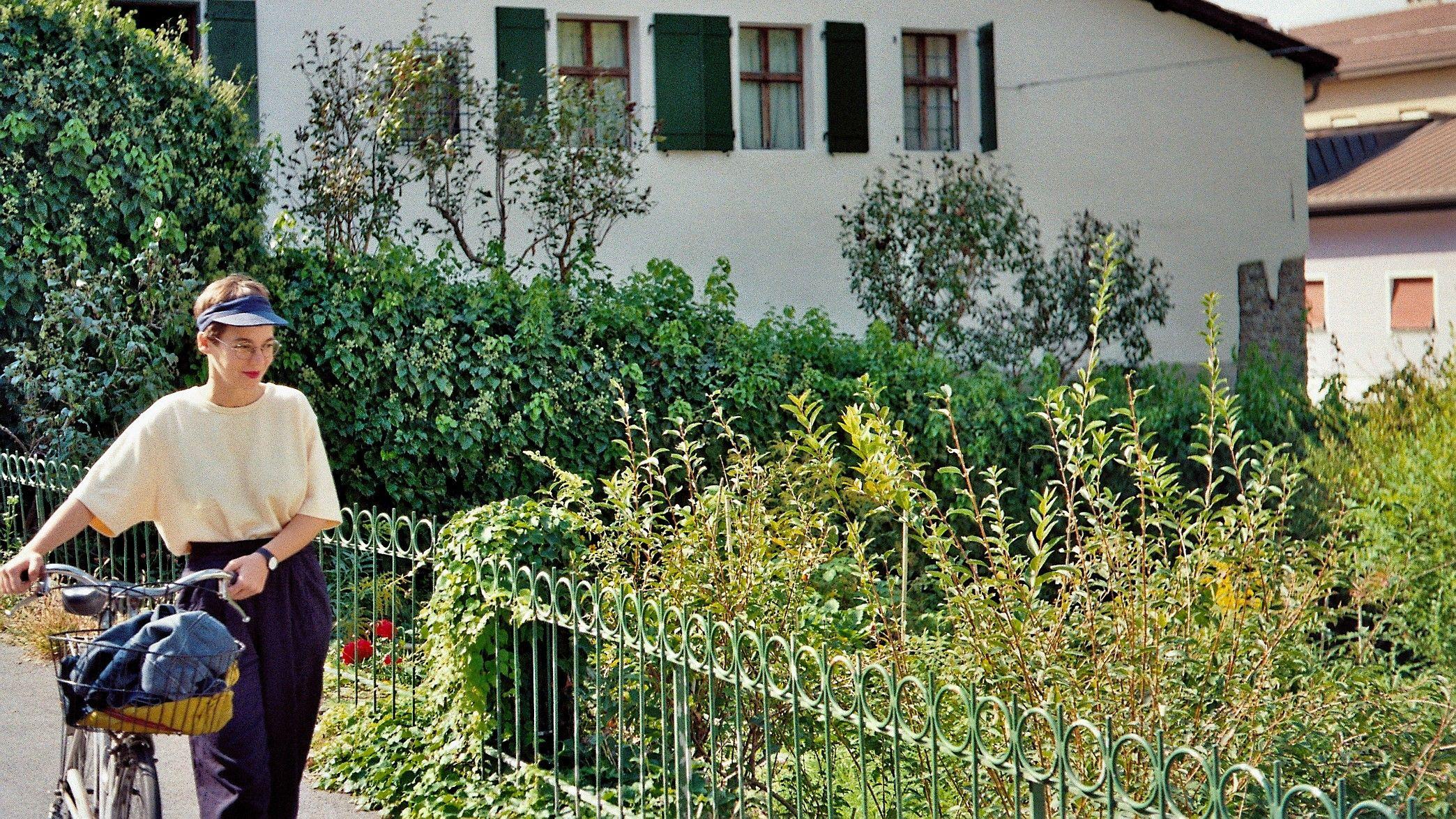A picture of Flora Mammana with a bicycle in front of a flourishing garden in the sun.