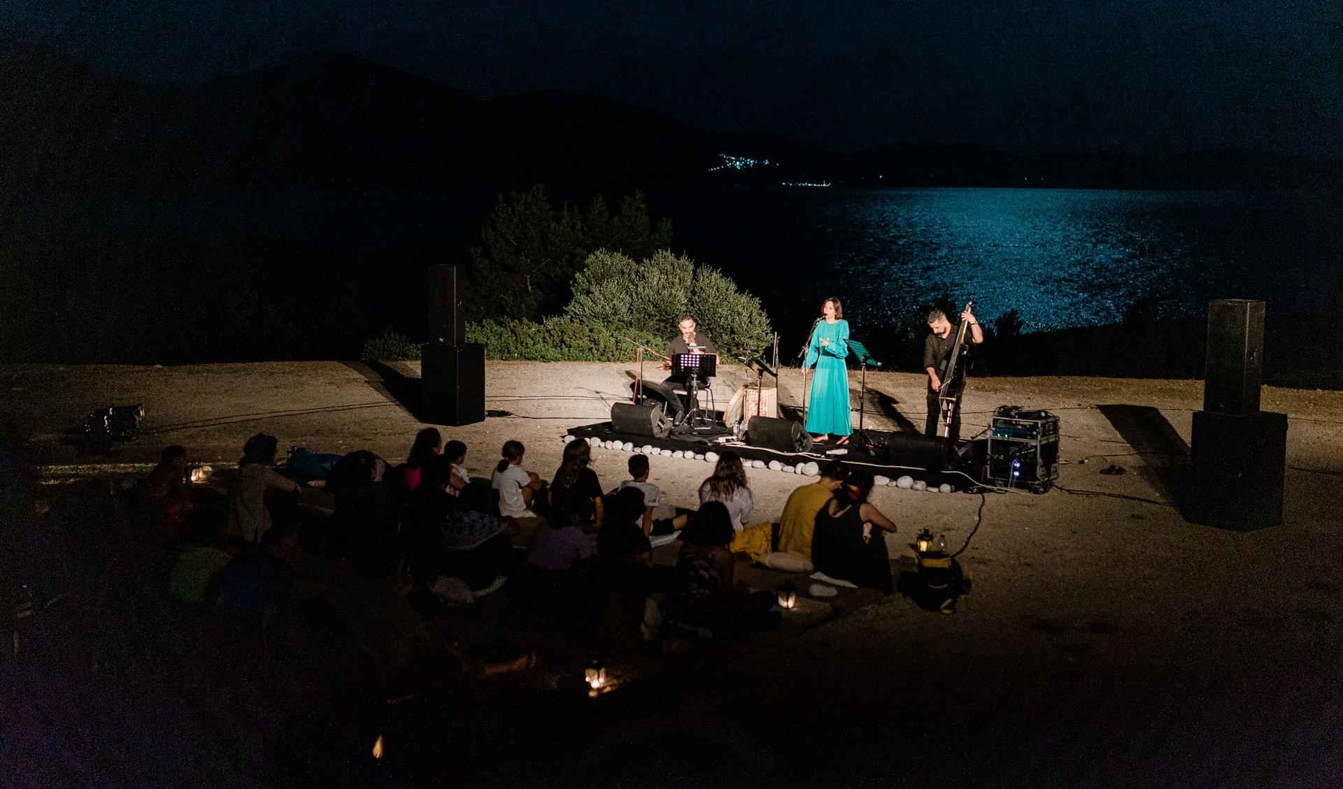 A small music performance: a women is singing and she is accomapnied by two musicians. In the background the sea.