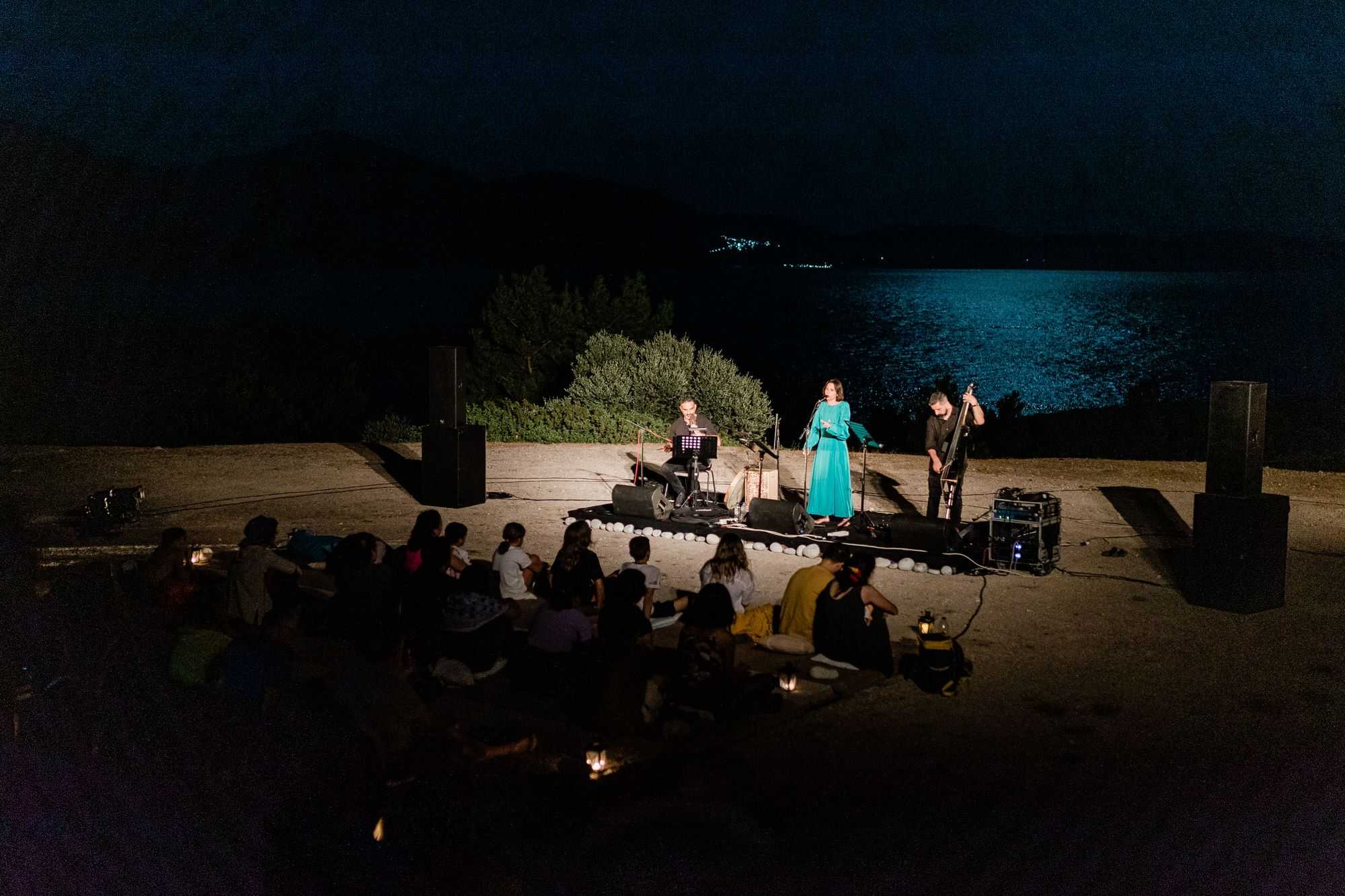 A small music performance: a women is singing and she is accomapnied by two musicians. In the background the sea.