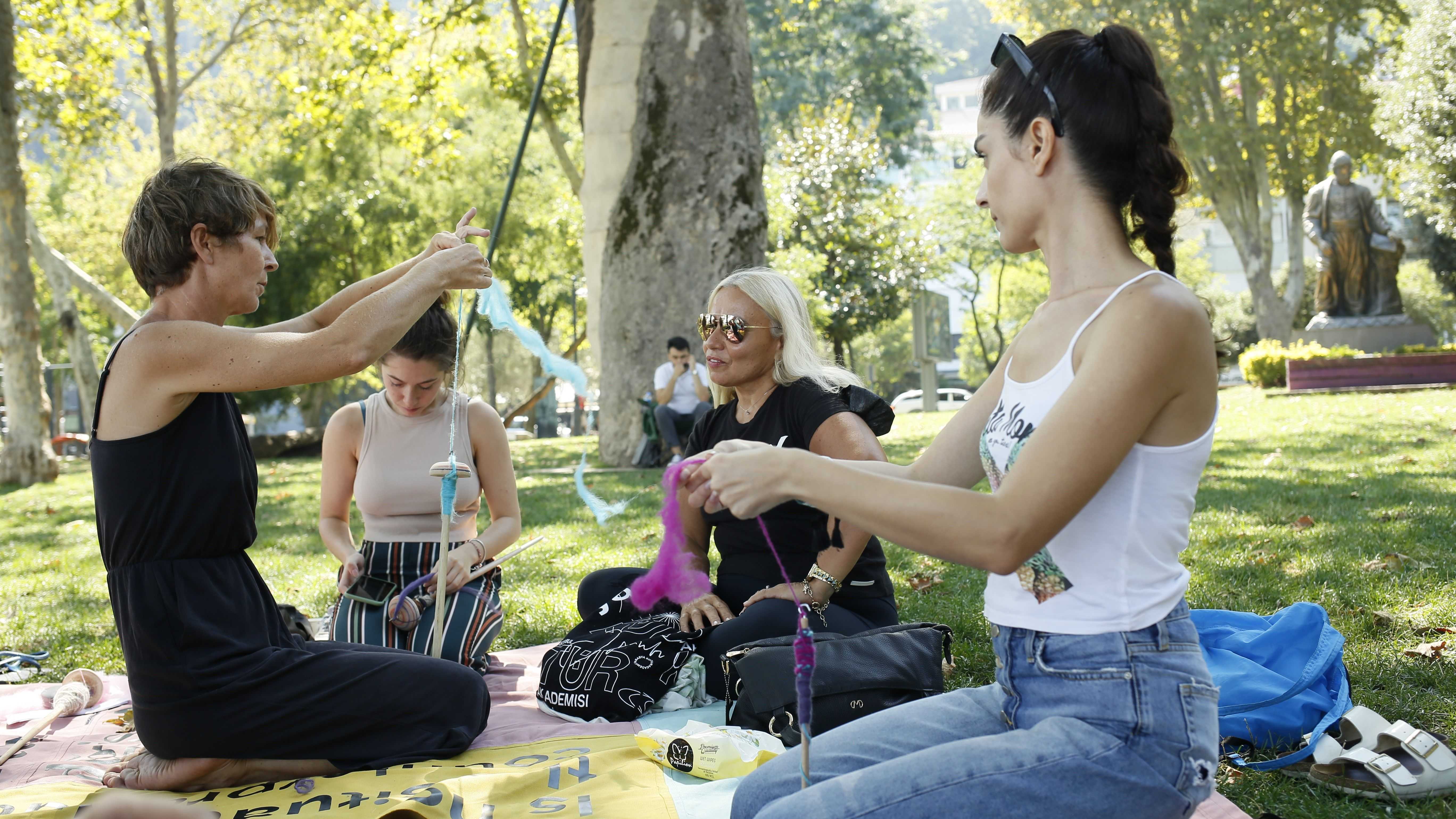 Four women are sitting in a park on a blanket and are watching and learning while one of them is showing them how to spin.
