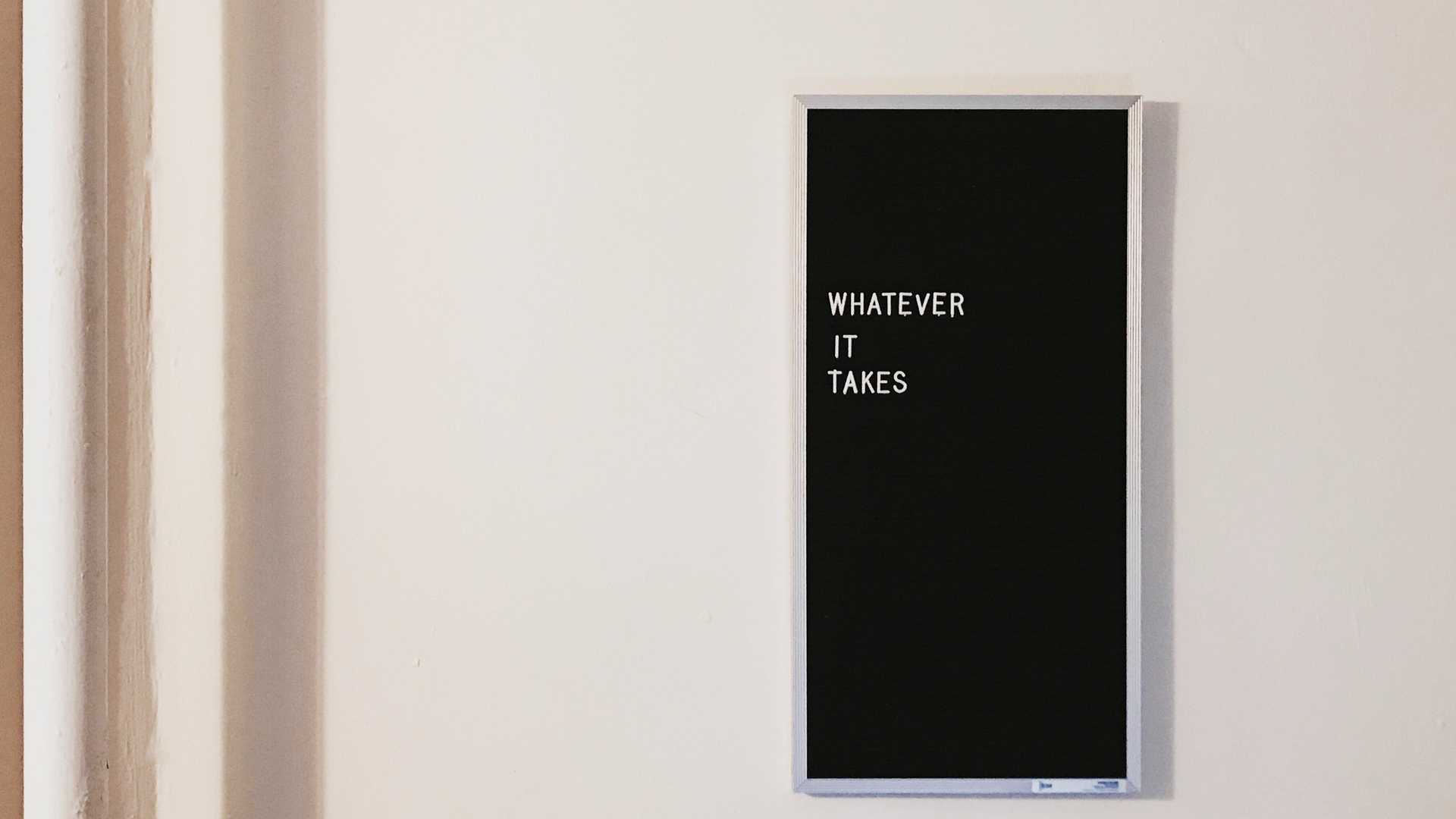 A white wall wih a black billboard. Its written on the billboard: Whatever it takes