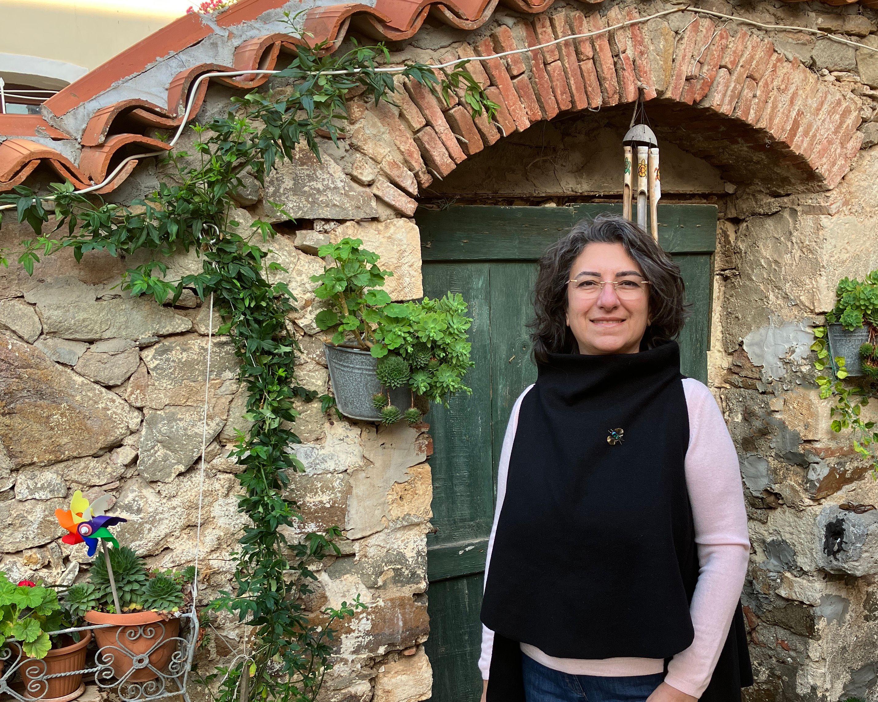 Ümit Hamlacıbaşı is standing in front of  a structure made of big stones. It looks like a wall. In the wall there is a green door and a wind chime. Ümit is smiling into the camera, is waering glasses and has long black hair.