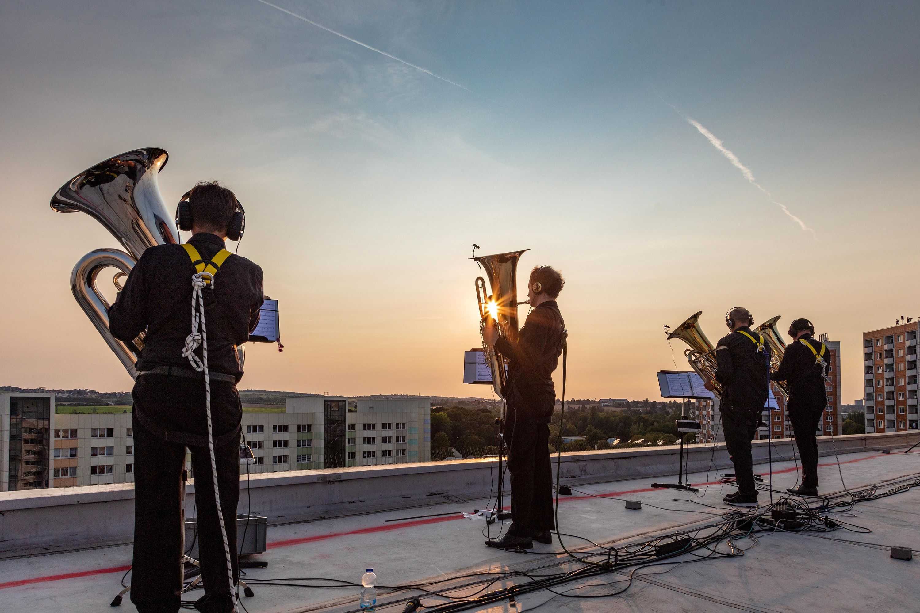 Four blowers, members of the Dresdner Sinfoniker are standing on the roof o a tower-builidng and are playing their instruments. They are secures with climbing gear and the sun goes down in the background