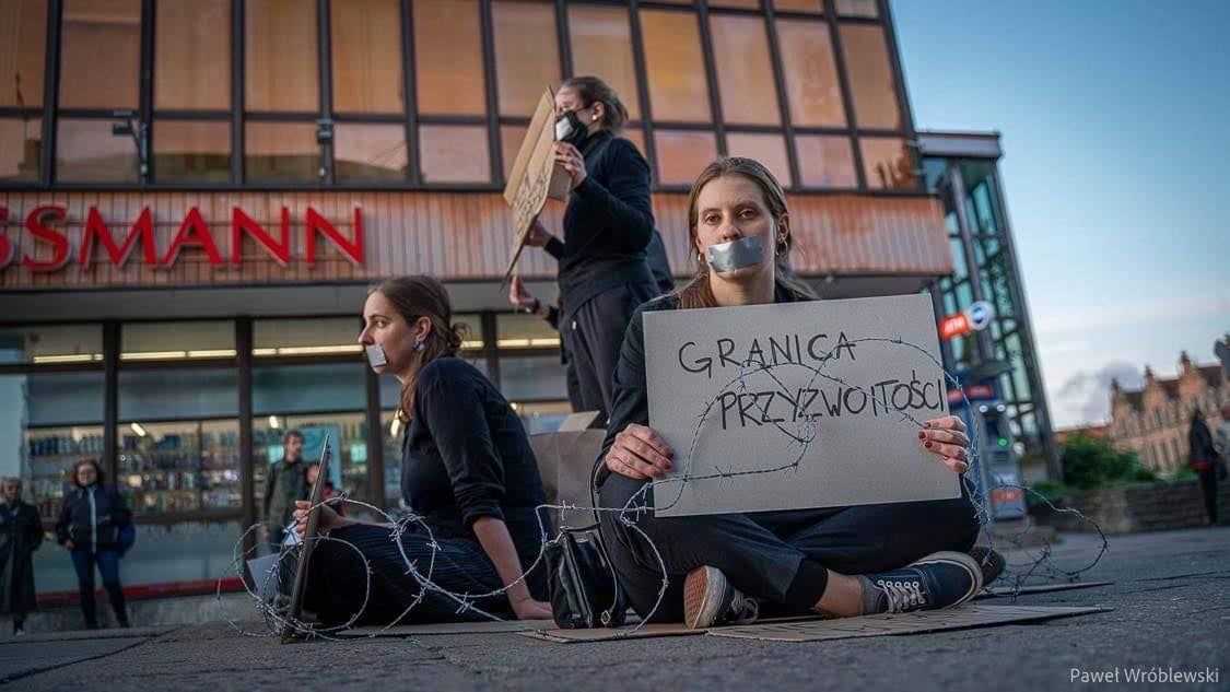Aleksandra Łoboda is sitting on a street, her mouth taped with gaffa and around her and two colleagues there is barbed wire. She is holding a sign that is saying: The border of Decency