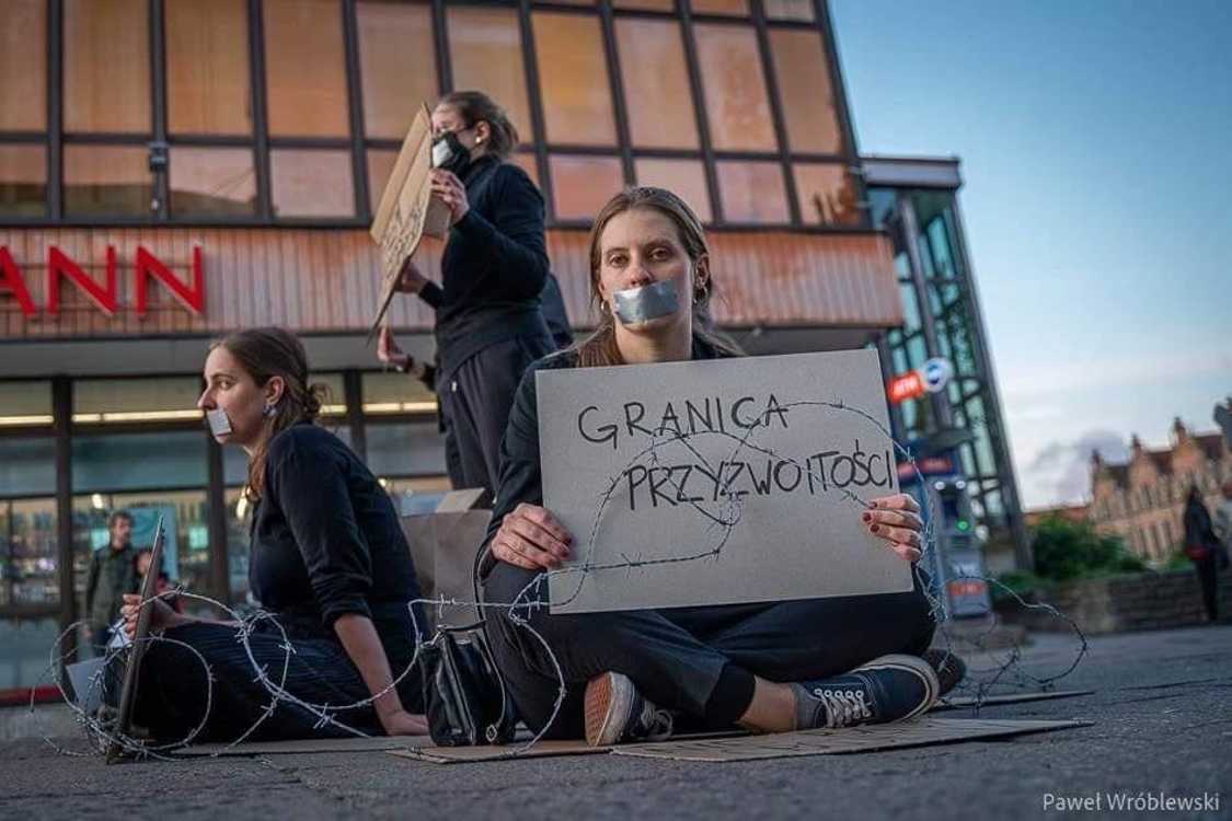 Aleksandra Łoboda is sitting on a street, her mouth taped with gaffa and around her and two colleagues there is barbed wire. She is holding a sign that is saying: The border of Decency