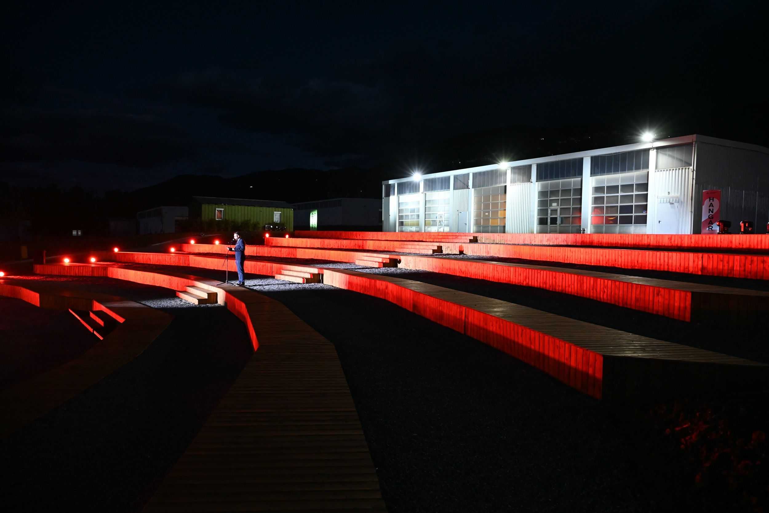 A person is standing, very well dressed in the front of a hangar. Its night time and the person is holding a speech.