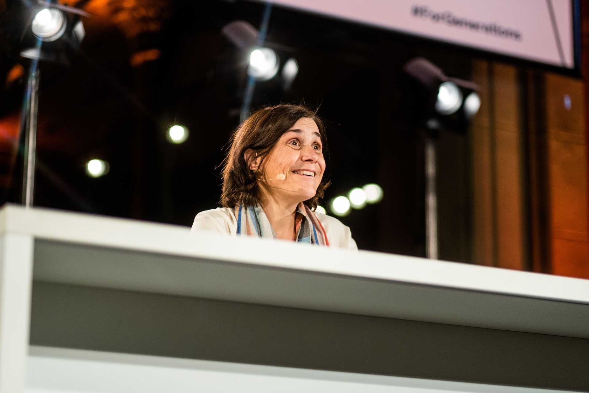 Bénédicte Savoy is leaning on a table and smiling towards the hall packed with people. Sheis smiling while she holds a speech. 